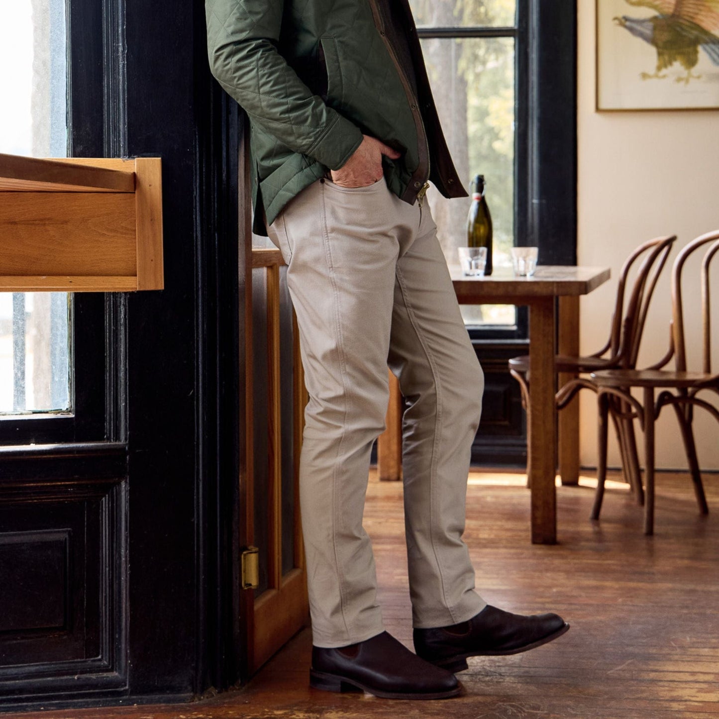 A person stands indoors in light-colored Tom Beckbe Redmont Pants and a green jacket, hands in pockets, beside a table and chairs.