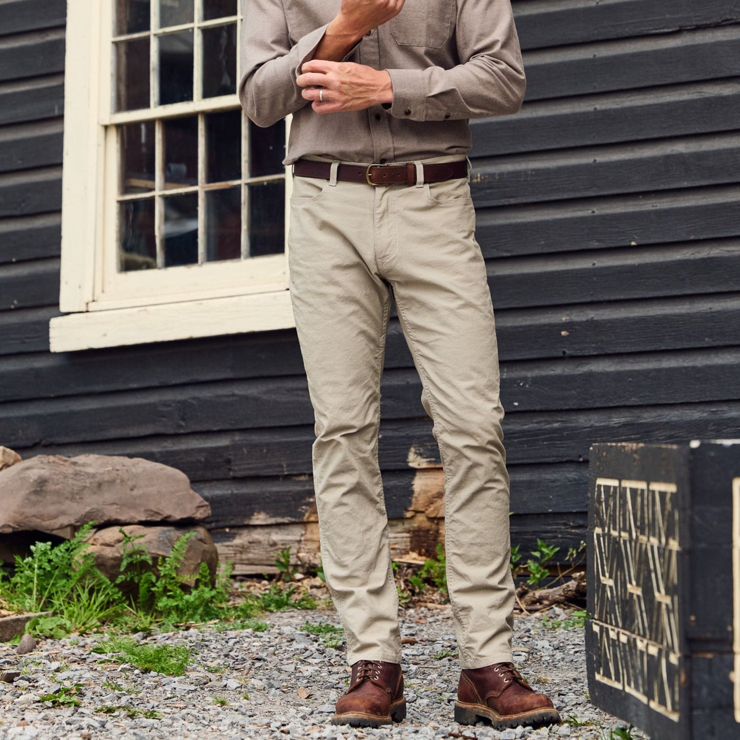 A person in a brown shirt and Tom Beckbe Redmont Pant stands outside by a wooden building and large crate.