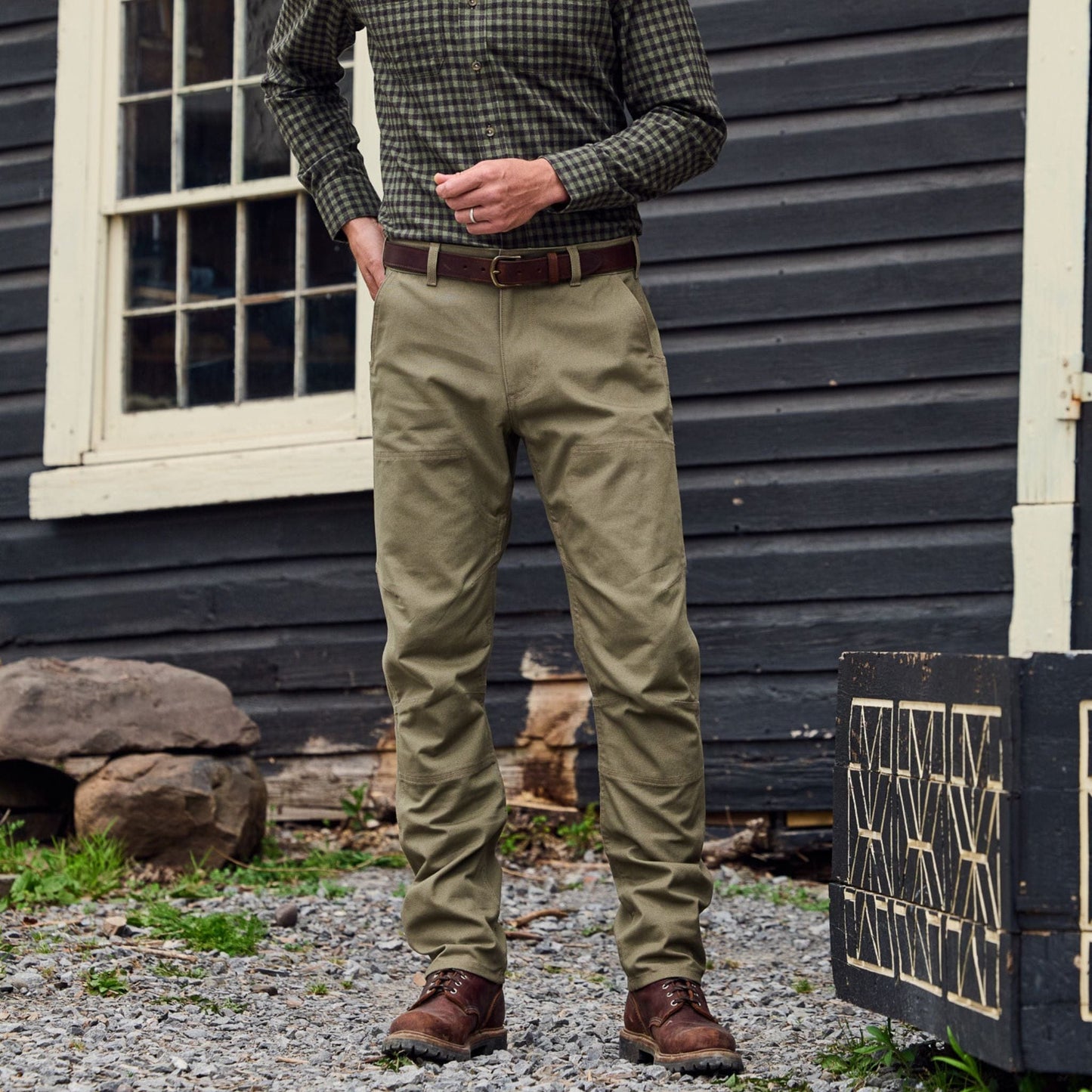 A person in a checkered shirt and Tom Beckbes Rutledge Double-Front Pant stands on gravel by a wooden box, with a dark house behind.