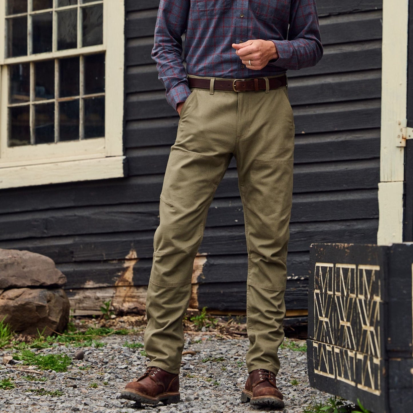 A man in a plaid shirt wearing Tom Beckbes Rutledge Double-Front Pant stands by a wooden building, hands in pockets.