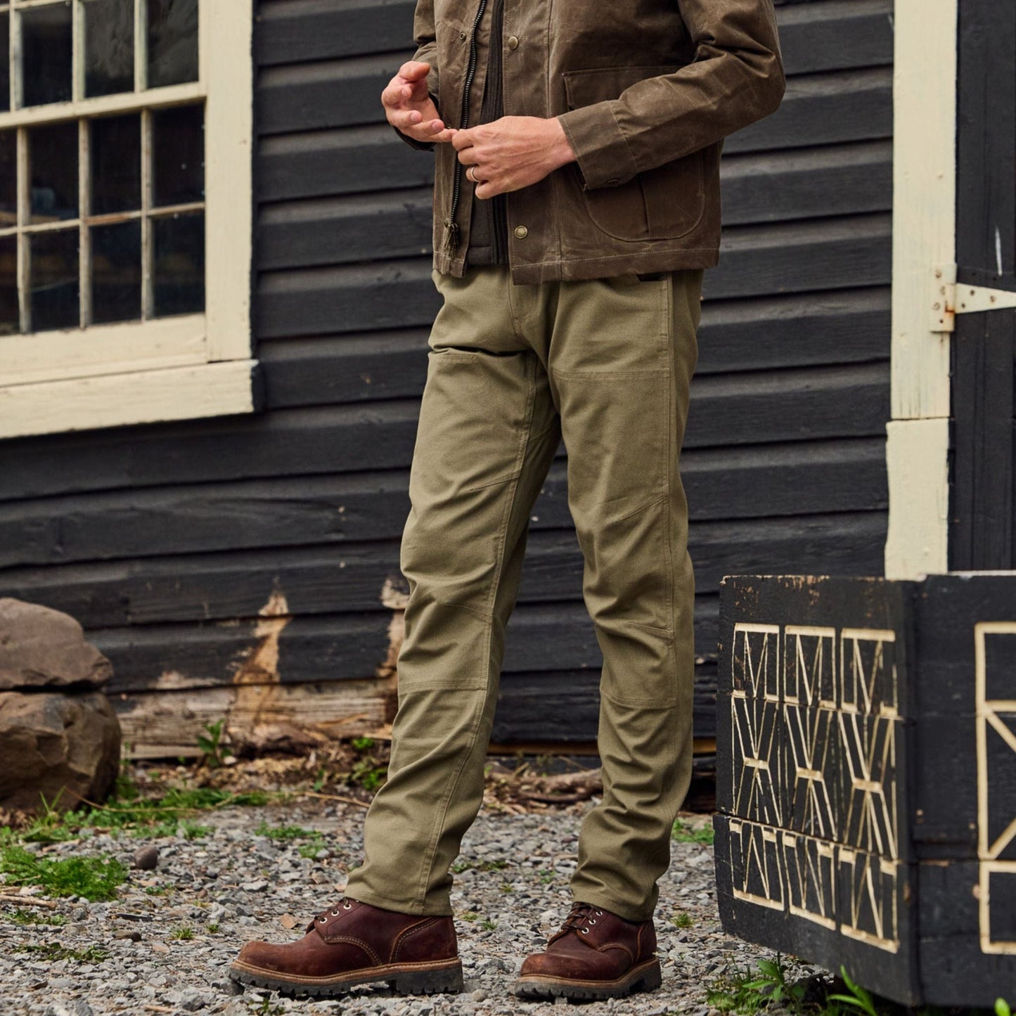 A person in a brown jacket and Rutledge Double-Front Pant stands outside a dark building near wooden boxes on gravel.
