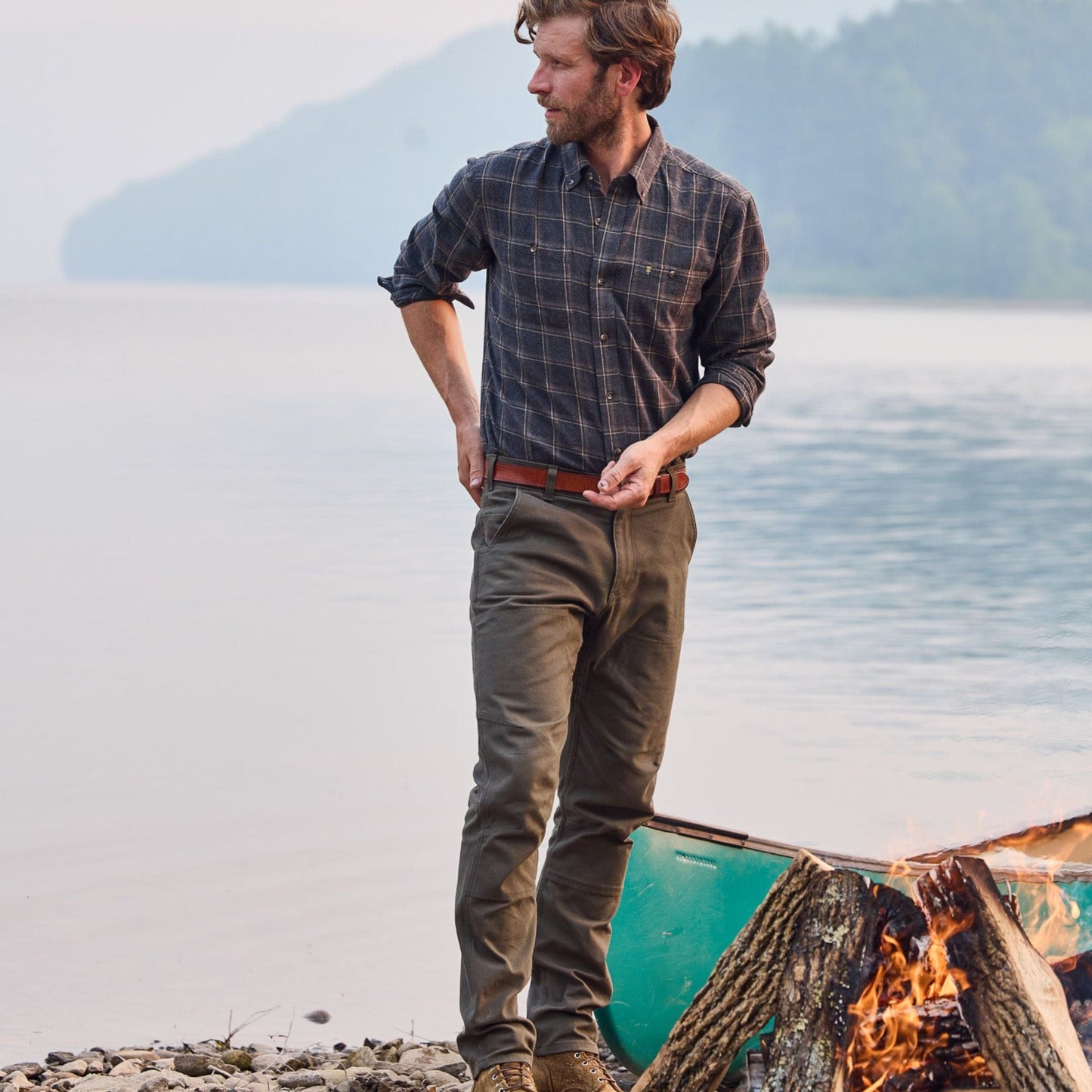 A man by a lakeside campfire in a checkered shirt and Tom Beckbe Rutledge Double-Front Pant, with a canoe behind.