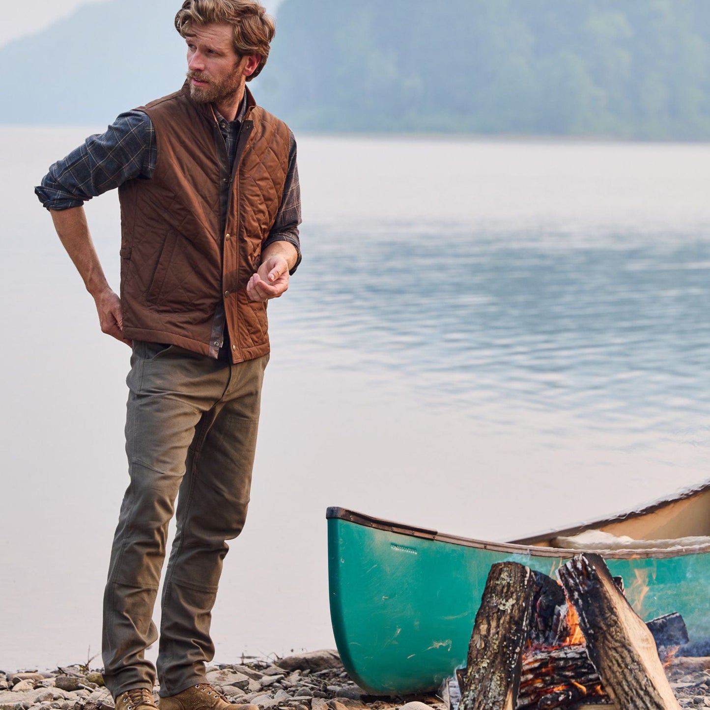 Man in Tom Beckbe Rutledge Double-Front Pant and plaid shirt by a canoe, with campfire, trees, and lake behind.
