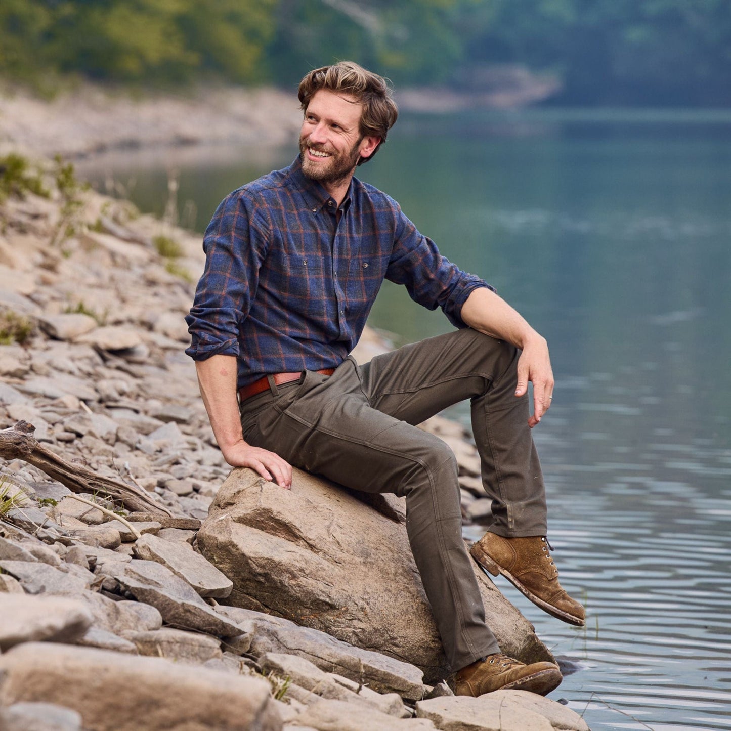 A man in a plaid shirt and classic fit Tom Beckbe Rutledge Double-Front Pant sits by a lake, surrounded by stones and trees.