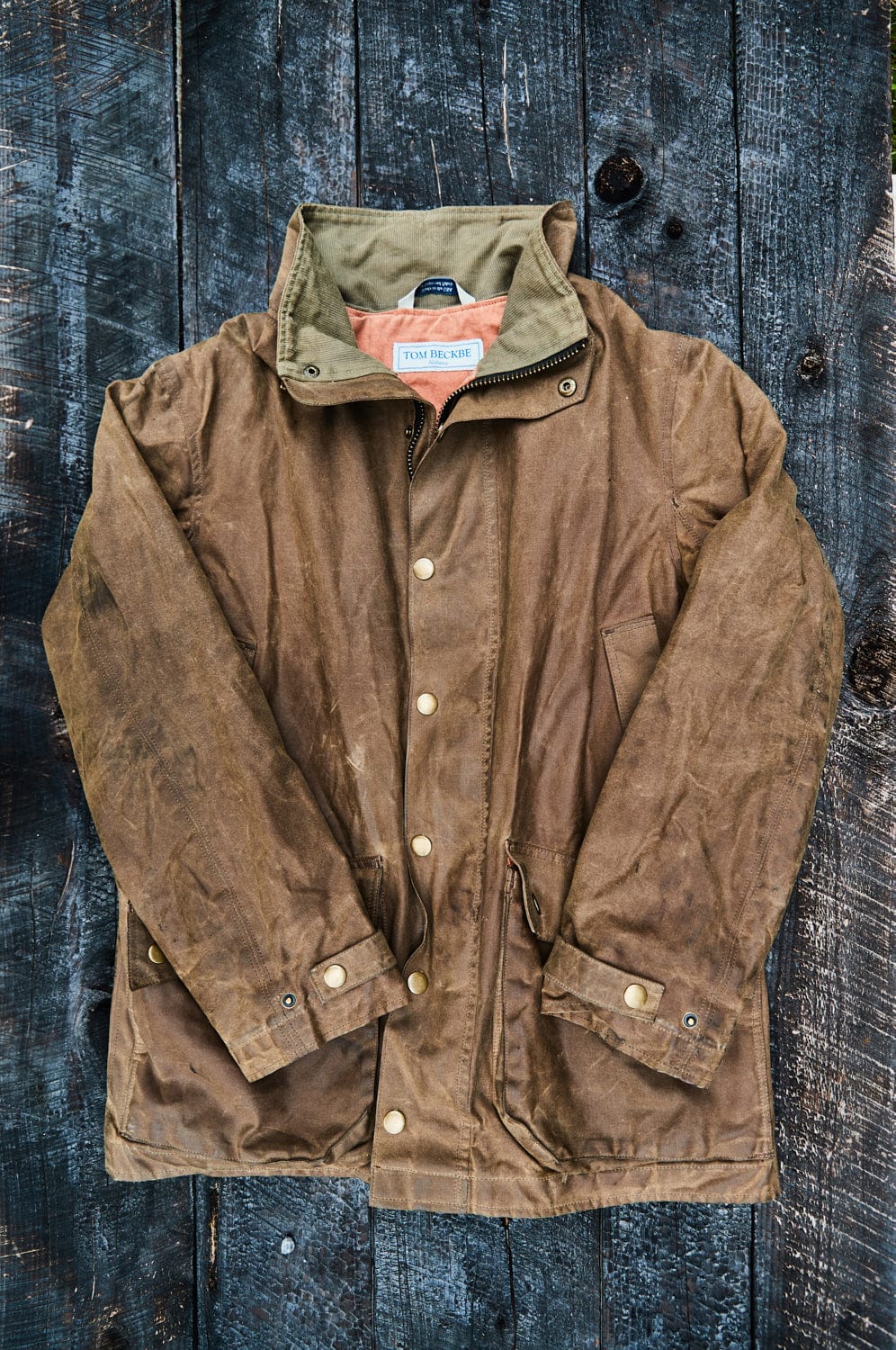 Brown waxed jacket with a collar and snap buttons, laid flat on a dark wooden surface.