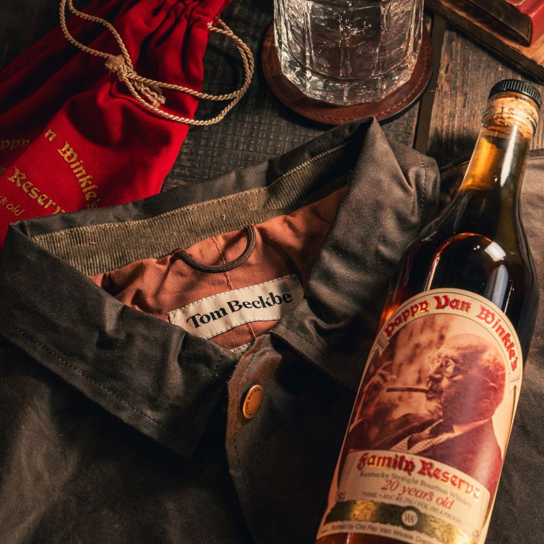 Bottle of Pappy Van Winkle bourbon, a dark jacket labeled Tom Beckbe, a red fabric bag, and a glass with clear liquid.