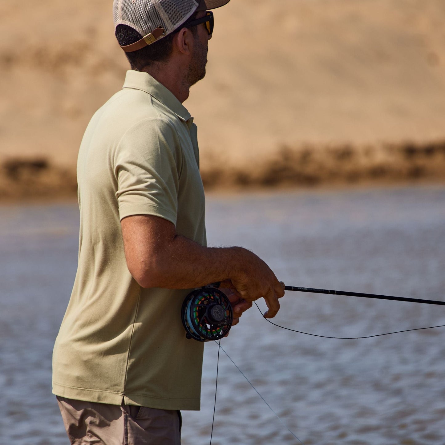 Man in a Tom Beckbe Coastal Polo fishing on a sandy beach, holding a rod with water and sand in the background.