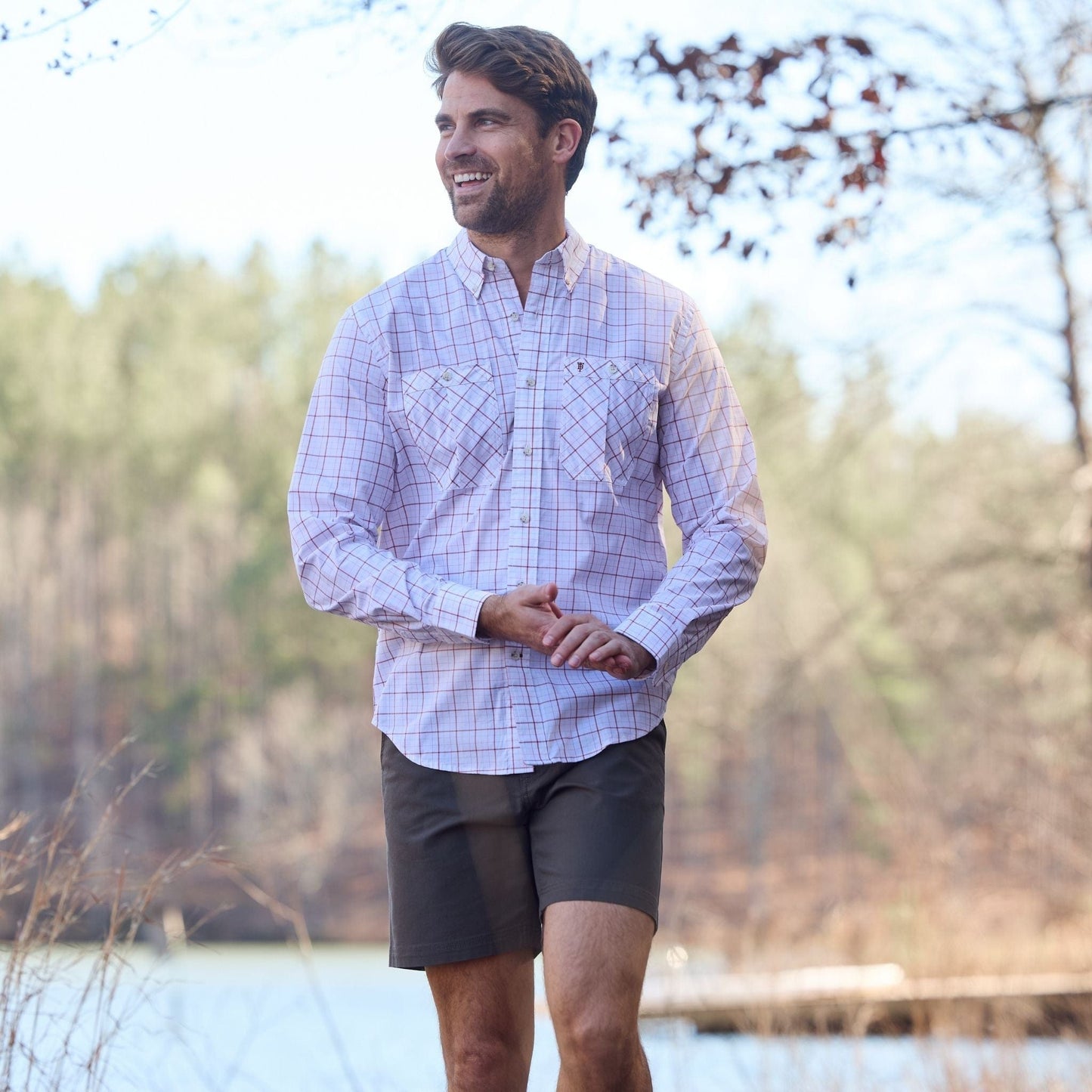 Man in a Tom Beckbe Tidewater Shirt smiles while walking near water and trees.