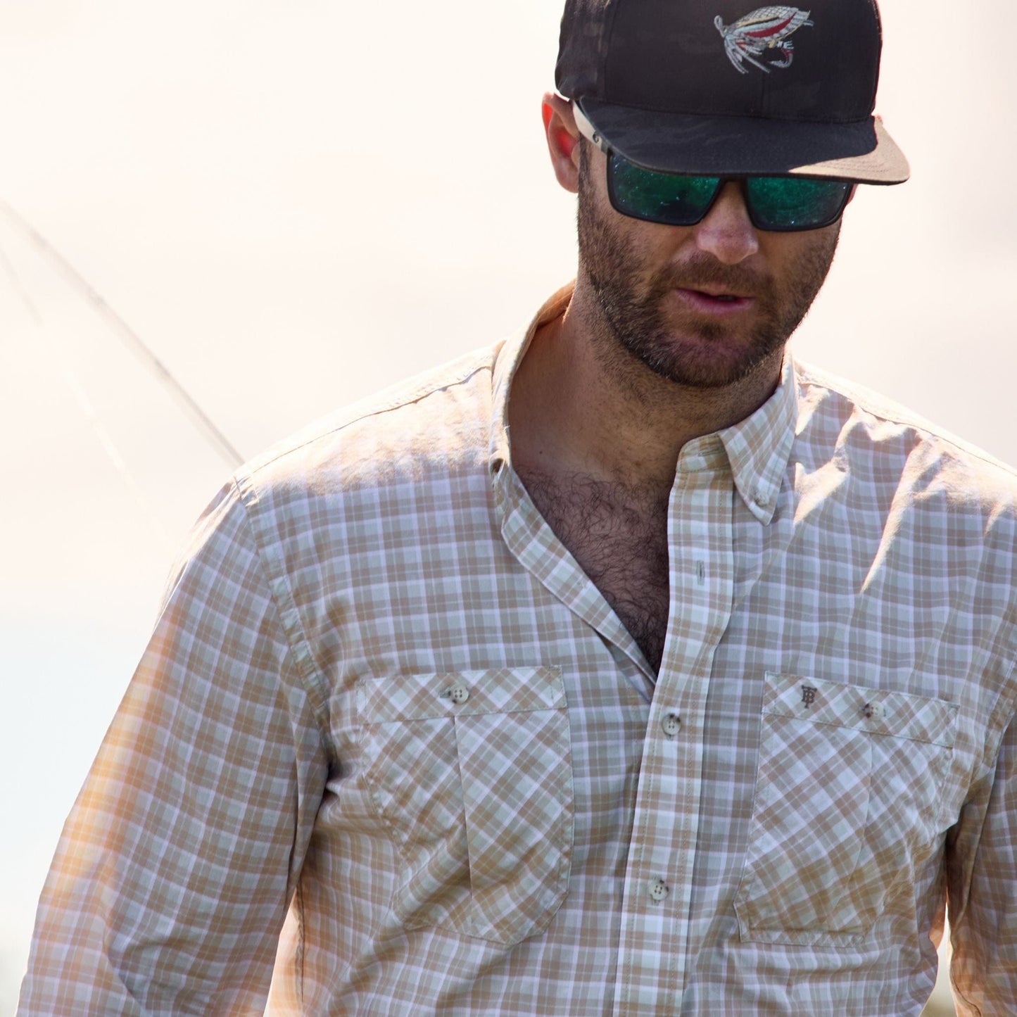 A man wearing a Tom Beckbe Tidewater Shirt, sunglasses, and black cap looks down against a neutral backdrop.