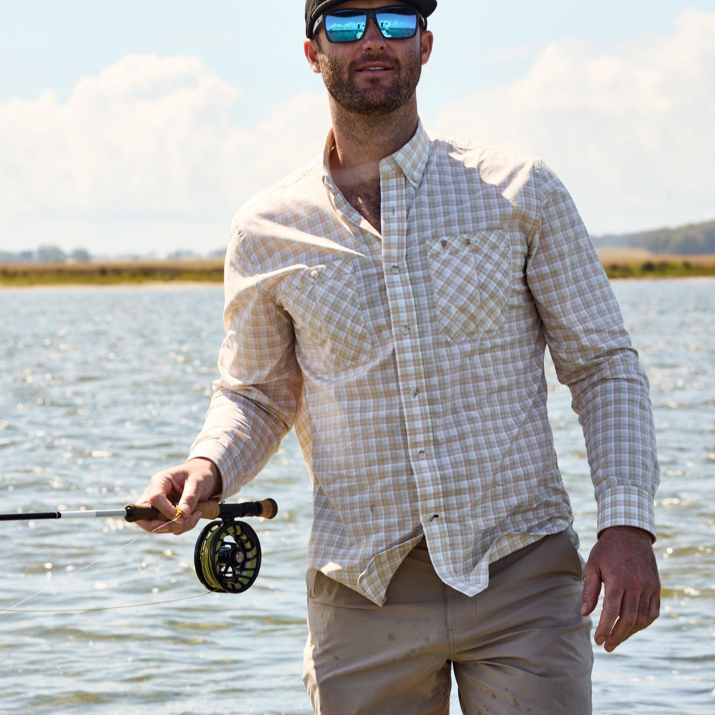 Person in water holding a rod, wearing a Tom Beckbe Tidewater Shirt and cap, against sky and water.