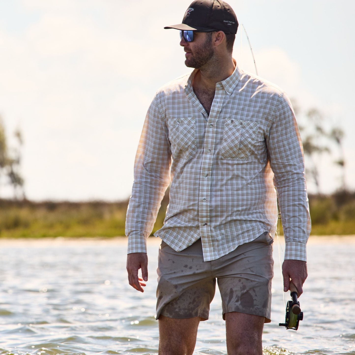 Man in shallow water with a fishing rod, wearing a Tom Beckbe Tidewater Shirt and cap, looking sideways.