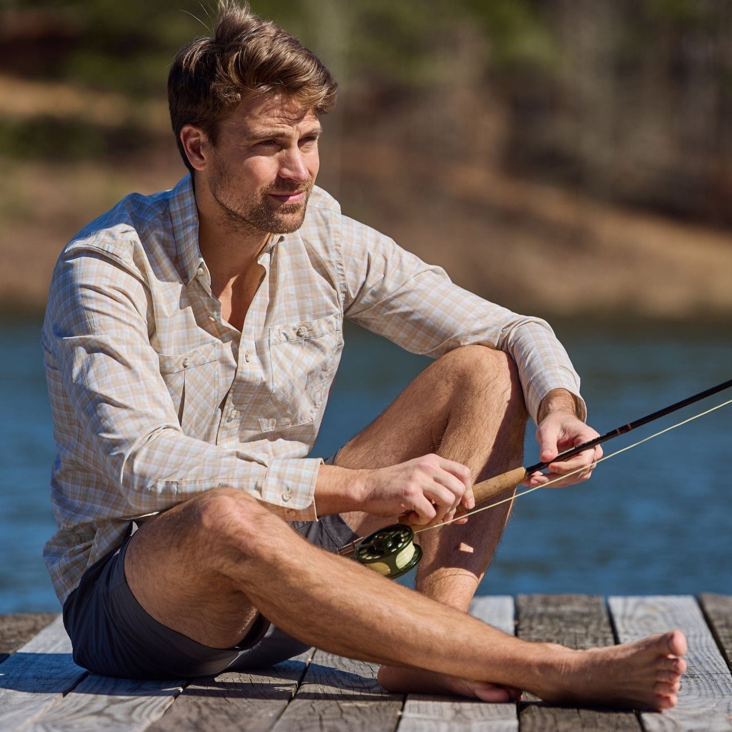 Man in a Tom Beckbe Tidewater Shirt is cross-legged on a dock, holding a fishing rod with water and trees behind him.