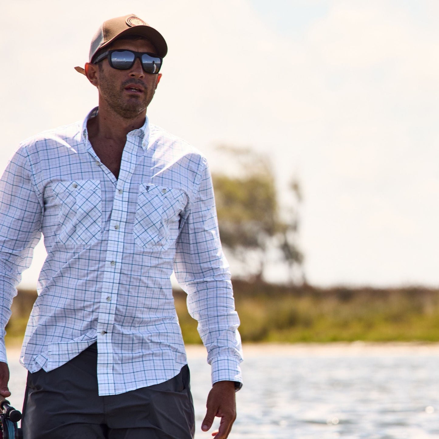 Man in a Tom Beckbe Tidewater Shirt, sunglasses, and cap stands by the water on a sunny day.