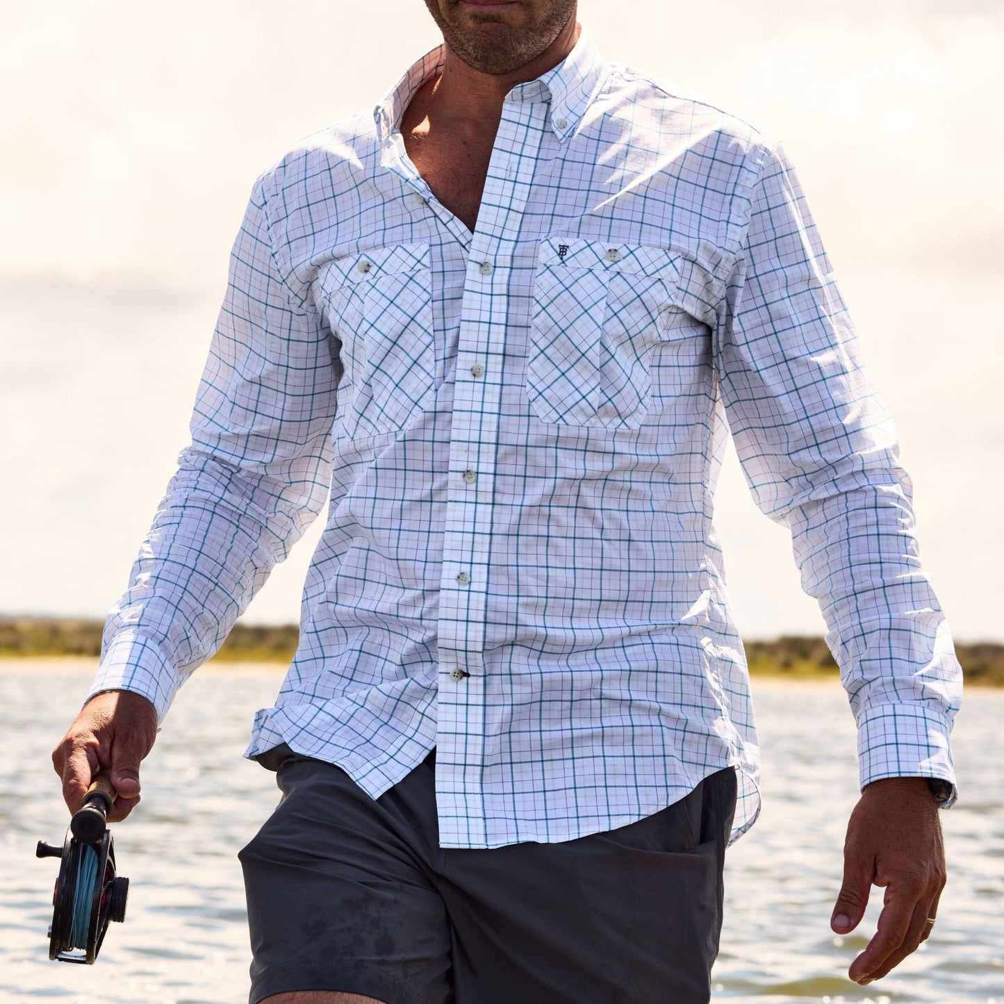 A man in a Tom Beckbe Tidewater Shirt stands in shallow water holding a fishing reel with UPF 30+ coverage.