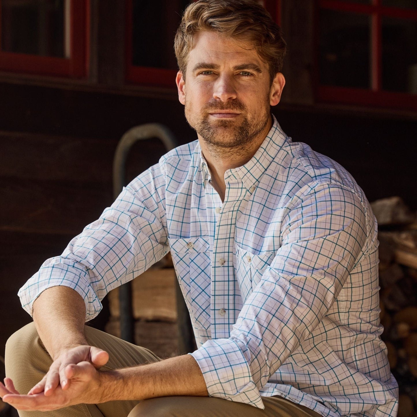 A bearded man in a Tom Beckbe Tidewater Shirt sits on steps before a wooden structure.