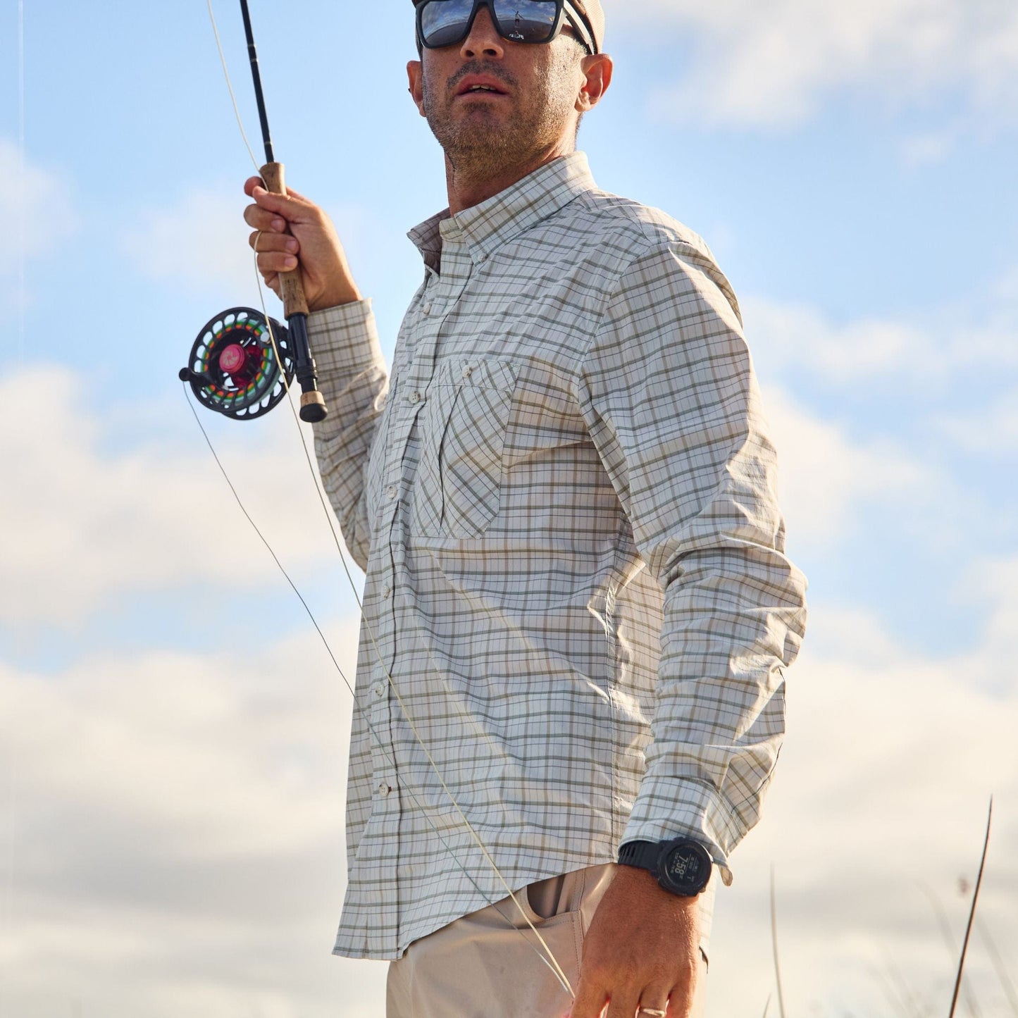 Man in sunglasses, wearing a Tom Beckbe Tidewater Shirt, holds a rod under a partly cloudy sky.