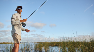 Man standing in shallow water among reeds, wearing a cap and sunglasses, holding a fishing rod, with blue sky and clouds in the background.