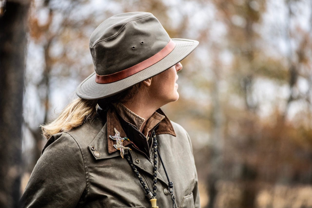 A person in outdoor clothing carries a shotgun over one shoulder and two dead birds in the other hand, walking through a grassy field with several birds in the foreground.