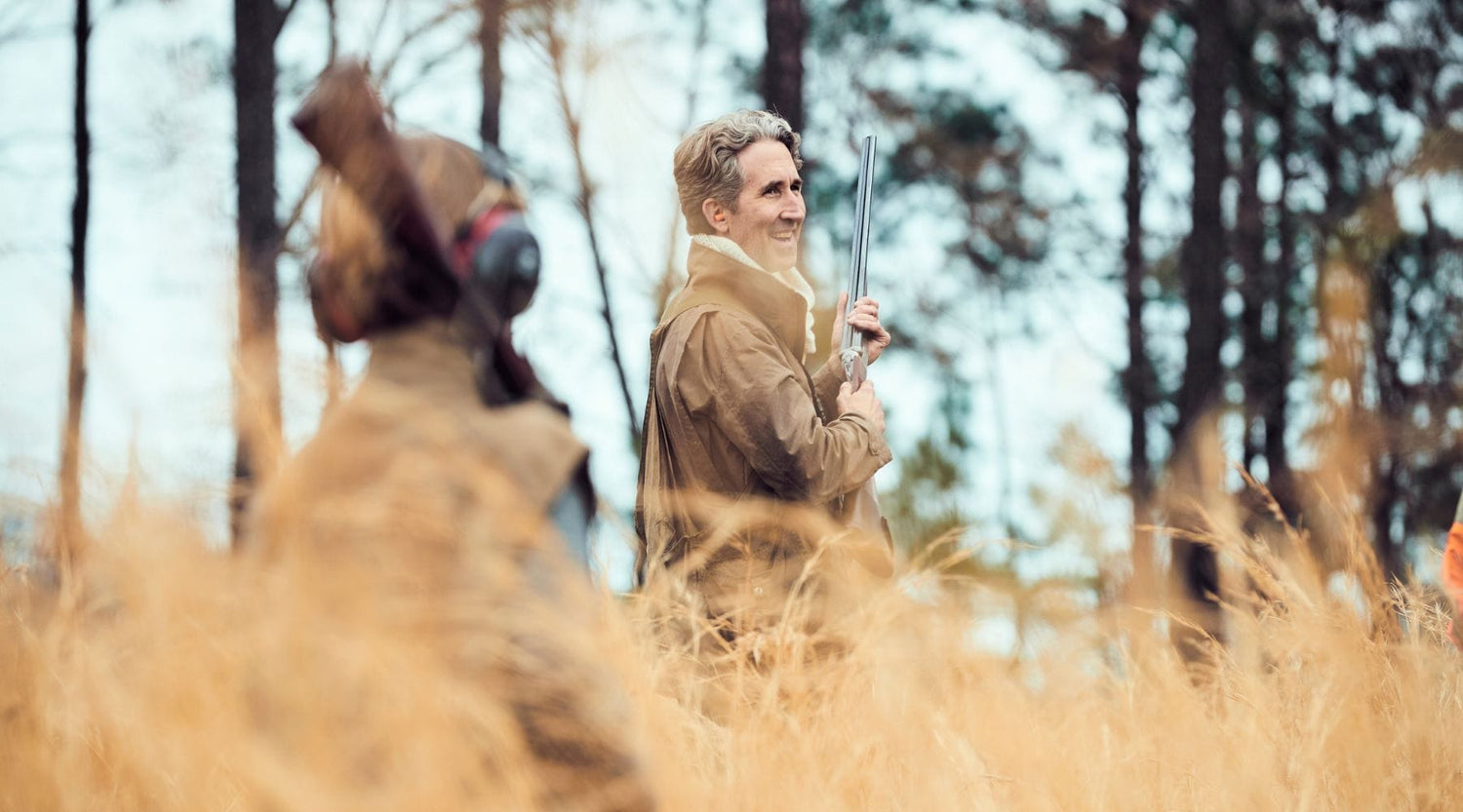 Two people dressed in brown outdoor clothing stand in tall grass, one holding a shotgun and smiling, with trees in the background.