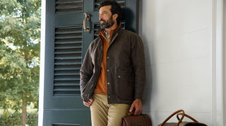 A man with a beard stands by a door, wearing a brown jacket over an orange sweater and khaki pants, with a brown leather bag placed beside him.