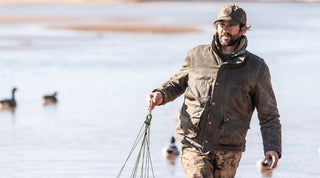 A man wearing an orange cap and vest walks through a forested area carrying a shotgun, surrounded by tall trees and dry grass.