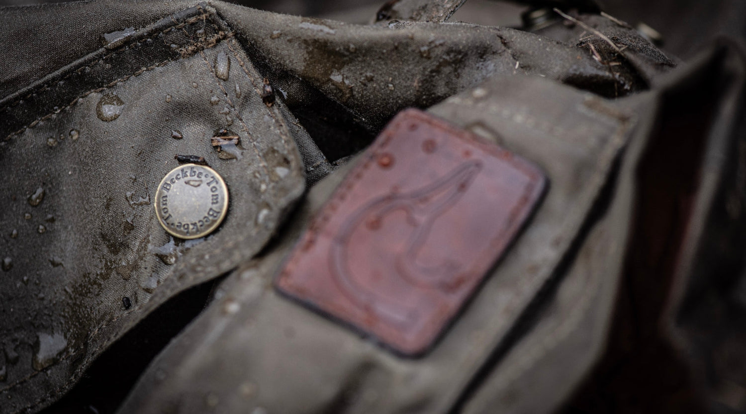 Close-up of a wet, brown fabric bag showing a leather logo patch and a round metal button with engraved text.