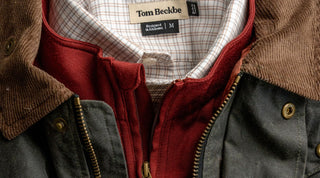A close-up of layered clothing: a dark jacket with brown corduroy collar, a red zip-up sweatshirt, and a white checkered shirt. The shirt label reads Tom Beckbe, Designed in Alabama, M..