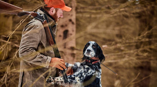 A man in hunting gear holding a shotgun stands beside a black and white dog in a wooded area during autumn.