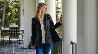 A woman with long blonde hair, wearing a dark jacket and blue jeans, stands on a porch holding a black bag and lightly touching a white column.