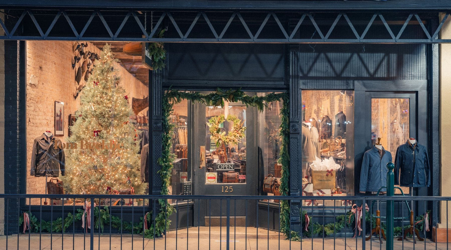 Clothing store with holiday decorations, including a lit Christmas tree and garlands, displays men’s apparel in the window, and an open sign on the door marked 125.