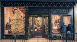 A clothing store decorated with Christmas garlands and a lit white Christmas tree, with mannequins dressed in winter apparel displayed in the front windows.