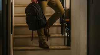 Person standing on stairs holding a black backpack in one hand, wearing olive pants and brown shoes, with only the lower body visible.