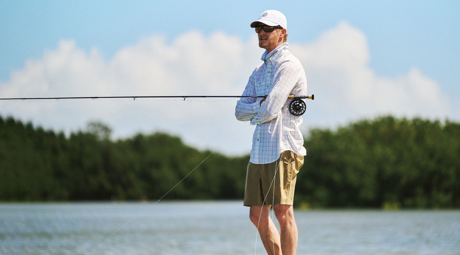 Man standing on a shoreline with arms crossed, holding a fishing rod, wearing sunglasses, a cap, and light-colored outdoor clothing. Trees and water are visible in the background.