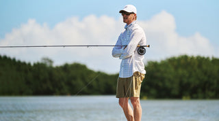 Man standing on a shoreline with arms crossed, holding a fishing rod, wearing sunglasses, a cap, and light-colored outdoor clothing. Trees and water are visible in the background.