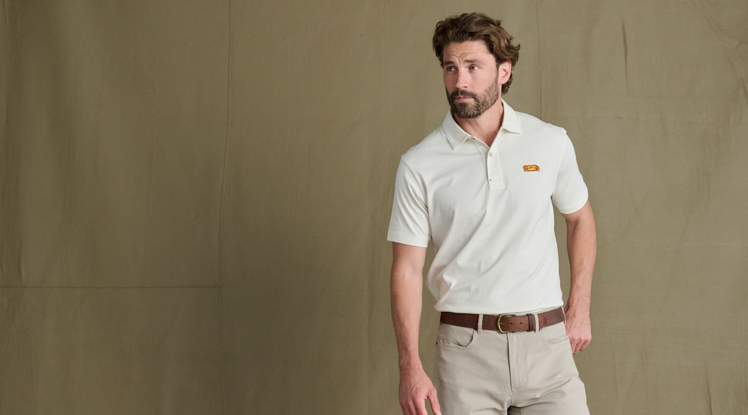 Man wearing a white polo shirt, beige pants, and a brown belt stands in front of a plain tan background.