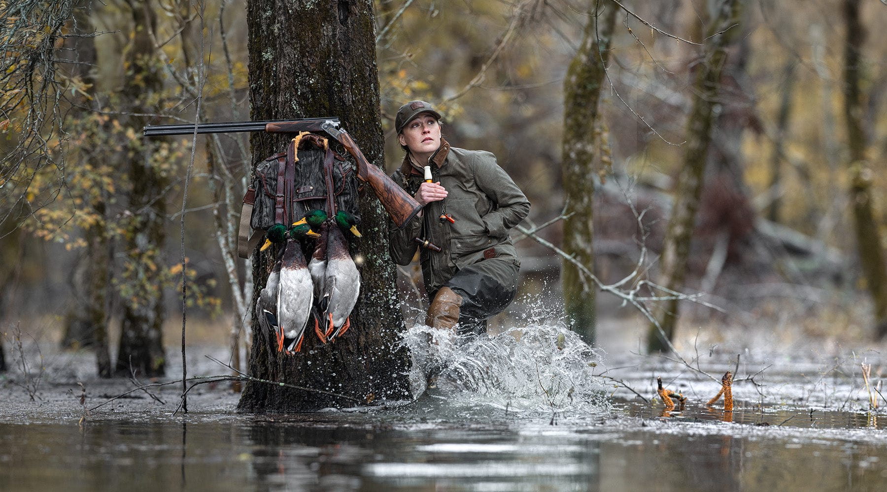 A person with a shotgun stands in flooded woods, leaning on a tree with ducks hanging from a backpack.