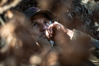 Man wearing a cap uses a duck call while partially concealed by dry branches and foliage.
