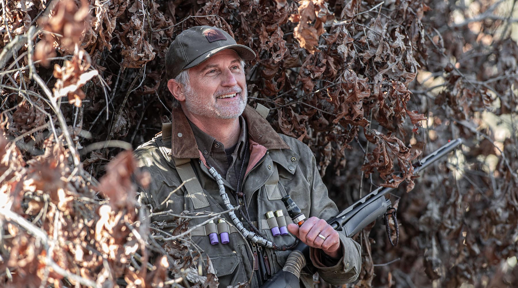 A man in outdoor gear holding a rifle stands in front of dried foliage, smiling and looking into the distance.