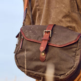Close-up of a Tom Beckbe canvas blind bag with leather straps and brass buckle, worn over a shoulder.