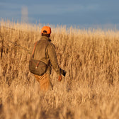 A person in an orange cap and jacket carries a shotgun and a Tom Beckbe Canvas Blind Bag through tall grass.