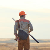 Wearing a hunting outfit, someone with a Tom Beckbe Canvas Blind Bag and rifle gazes at the sunset in a flat landscape.
