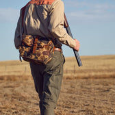 Person in outdoor attire carrying a shotgun and Tom Beckbes Classic Camo Blind Bag, walking in a field under blue skies.