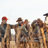 Group of men in a field wearing waxed canvas jackets and hats make a toast after a hunt with a dog.
