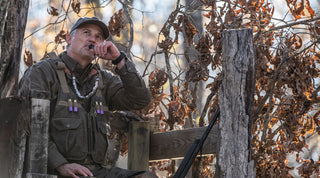 A person in outdoor gear uses a duck call while seated in a wooden blind with a shotgun beside them in an autumn forest.