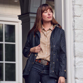 A woman in a dark jacket and checked shirt stands by a white brick wall, wearing Tom Beckbe's Crosshatch Quail Belt Buckle.