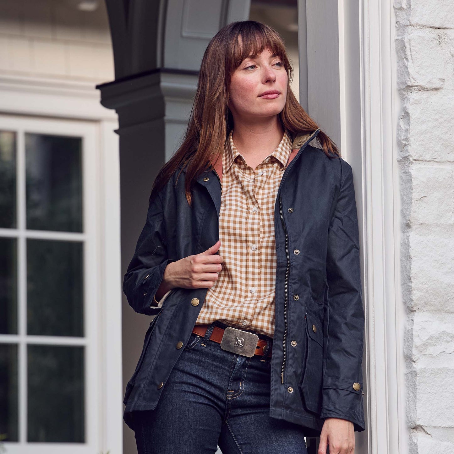 A woman in a dark jacket and checked shirt stands by a white brick wall, wearing Tom Beckbe's Crosshatch Quail Belt Buckle.