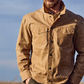 A man wears a Tom Beckbe Chatom Jacket, collar up, outdoors with hands in pockets against a clear sky.