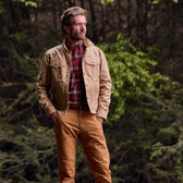 A man stands outdoors in a Tom Beckbe Chatom Jacket, red plaid shirt, and brown pants, hands in pockets, surrounded by trees.