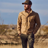 A man in a Tom Beckbe Chatom Jacket and cap walks outdoors in a dry, blurred field.