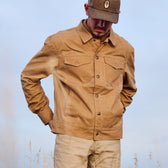 Man in a Tom Beckbe Chatom Jacket and hat, looking down with hands in pockets, stands outside under a clear sky.